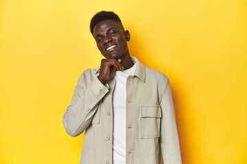 Stylish young African man on vibrant yellow studio background, smiling happy and confident, touching chin with hand.