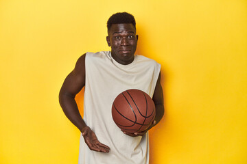 Athletic African youth posing with basketball, yellow backdrop, shrugs shoulders and open eyes...