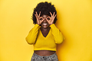 African-American woman with afro, studio yellow background showing okay sign over eyes