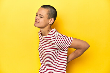 Shaved head woman in red striped tee, yellow backdrop suffering a back pain.