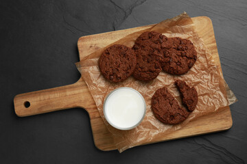 Board with tasty chocolate cookies and glass of milk on dark table, top view