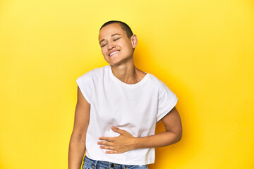 Shaved head woman in white tank top, yellow backdrop touches tummy, smiles gently, eating and satisfaction concept.