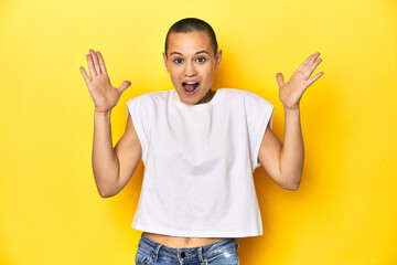 Shaved head woman in white tank top, yellow backdrop celebrating a victory or success, he is surprised and shocked.