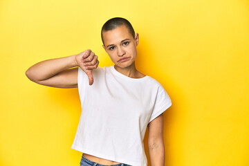 Shaved head woman in white tank top, yellow backdrop showing a dislike gesture, thumbs down. Disagreement concept.