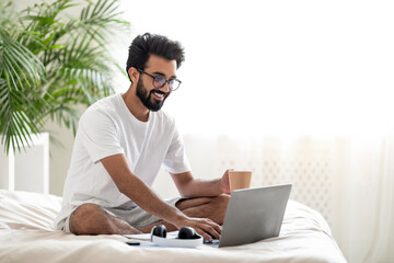 Young Indian Man Using Laptop And Drinking Coffee While Sitting In Bed