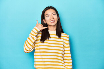 Asian woman in striped yellow sweater, showing a mobile phone call gesture with fingers.