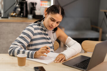 Online education concept. Female student studying remotely using a laptop, listening online lesson, taking notes in a notebook, while sitting in a coffee shop.