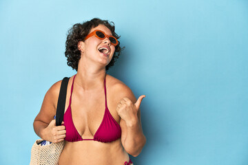 Young woman in bikini with beach bag points with thumb finger away, laughing and carefree.