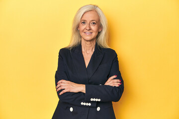 Confident middle-aged woman in business jacket, arms crossed, yellow studio.