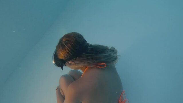 Cinematic shot of a woman in an orange bikini and vintage diving goggles moving gracefully underwater in clear blue waters shot in 4K, 120 FPS, Slomo