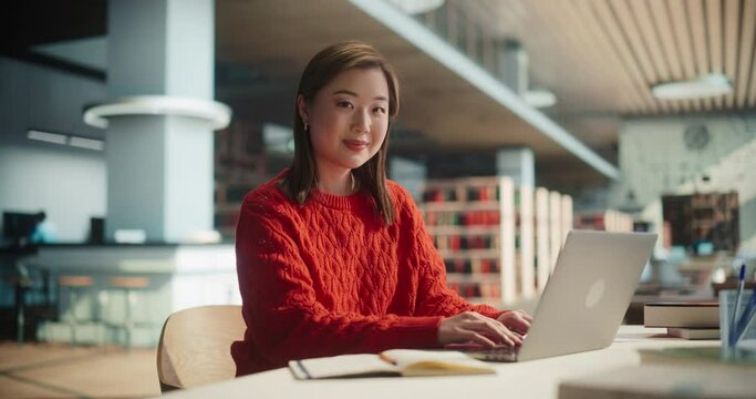 Beautiful Japanese Female Student Working on Her College Degree Thesis on a Laptop Computer in Library. Young Asian Woman Looking at Camera, Smiling. Girl Wearing a Casual Red Jumper