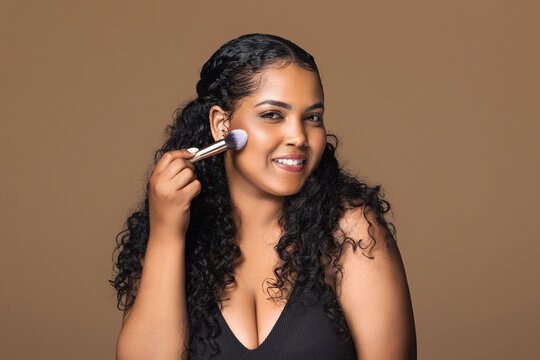 Cheerful latin chubby lady with makeup brush applying blush on her cheeks, enjoying brand new makeup products