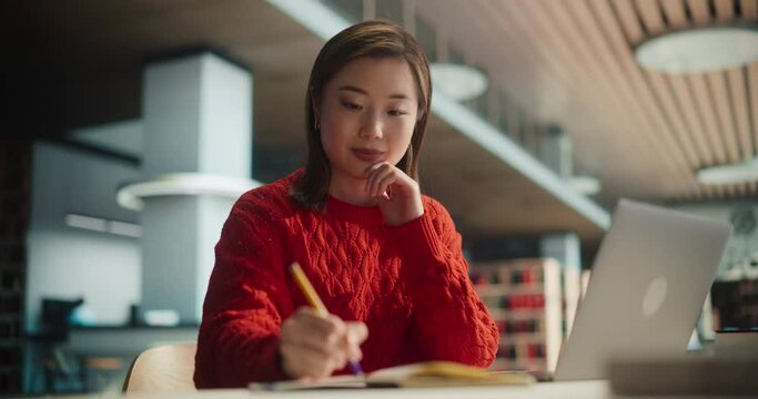 Attractive Multiethnic Student Making a Communication and Journalism Class Homework in a Textbook, While Researching Answers on a Laptop Computer. Young Asian Female Studying in a Public Library