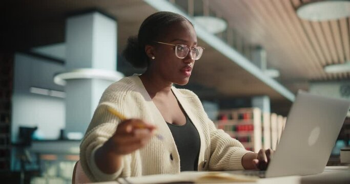 Portrait of a Stylish African Female Student in Glasses Working on Her University Exercise on a Laptop Computer. Young Black Woman Studying an Online Course in an Empty Quiet Public Library
