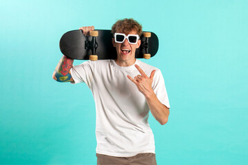 young cheerful tattooed guy in white t-shirt holds skateboard and shouts on blue isolated background