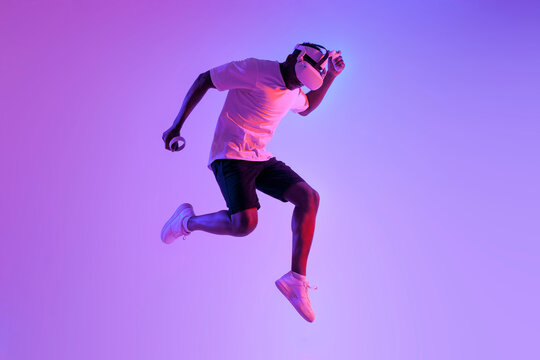african american athletic man jumping in virtual reality glasses in neon lighting, the guy athlete runs and trains