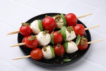 Caprese skewers with tomatoes, mozzarella balls, basil and pesto sauce on white tiled table, closeup