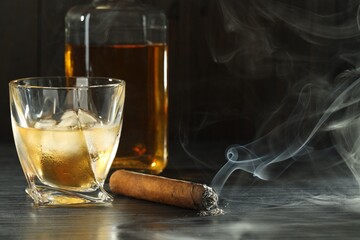 Bottle, glass of whiskey with ice cubes and smoldering cigar on black wooden table. Space for text
