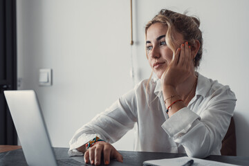 Thoughtful millennial student girl sitting at table with laptop, open books, looking away, thinking...