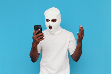 shocked african american male hacker in white balaclava uses smartphone and is surprised on blue isolated background