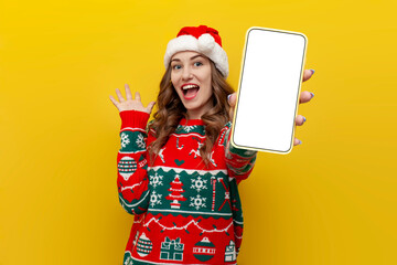 girl in christmas sweater and santa claus hat shows blank smartphone screen on yellow isolated...