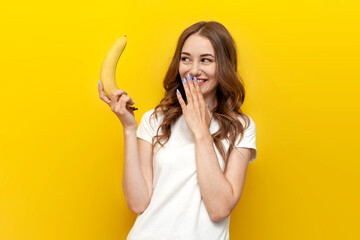 young girl holding small banana and mocking on yellow isolated background, woman presents erotic...