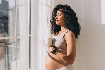 Charming lovely pregnant female in crop top patting her belly standing in front of window under sun rays, drinking coffee or tea in morning, talking too her baby, imagining their future life together - Powered by Adobe