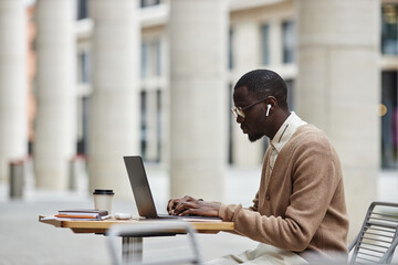 Side view of young African American businessman in earphones and casualwear sitting by table in...