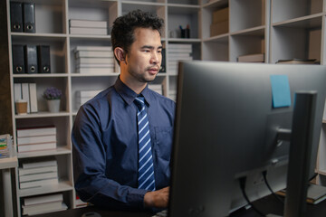 Asian man sitting in the office working overtime in the middle of the night, employee working overtime to get work done on time. The concept of overtime and hard work of company employees.