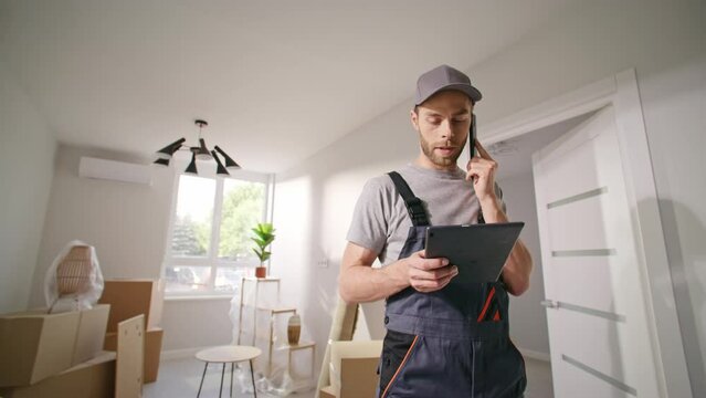 Master came to make repair work. Builder in special overalls in flat. Contractor talking by phone with boss. Repairman communicating with foreman. Man holding folder on background of ladder and boxes.