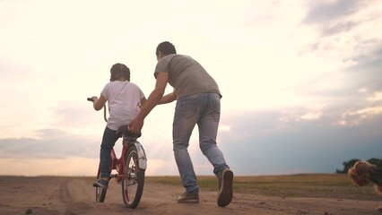 dad teaches son to ride a bike. happy family kid dream concept. the boy sat on bicycle for the first time, his father teaches boy to ride a bicycle. dog lifestyle runs with family, fun family pastime