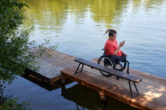High angle portrait of young man with disability sitting in wheelchair by lake and using smartphone, copy space