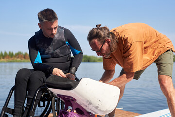 Portrait of senior sports instructor helping man with disability and assembling adaptive equipment for wakeboarding