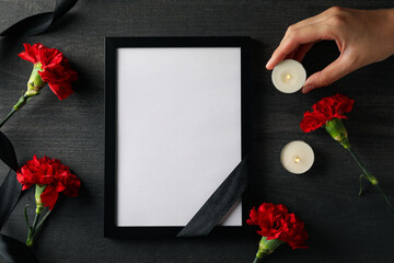 Flowers, photo frame, black ribbon and candles in female hand on black background, top view