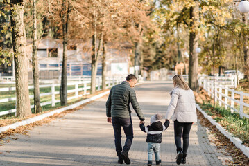 Mom, dad hold hands son child walking in park at sunset. Family spending time together on vacation. Mother, father, kid running in street near forest in nature. Concept of autumn holiday. Back view.