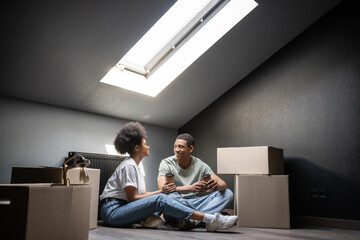 joyful african american couple with coffee to go talking near carton boxes on attic in new house