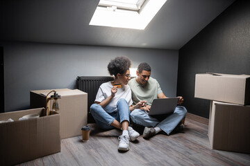 Fototapeta na wymiar smiling african american couple using laptop and credit card near boxes on attic in new house