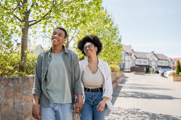 positive african american couple holding hands while walking on urban street in summer