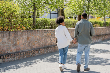 Obraz premium smiling african american woman holding hand of boyfriend while walking on urban street