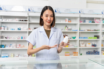 Happy beautiful asian female pharmacist wearing lab coat standing and holding pill bottle, She...