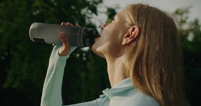 Closeup of beautiful young woman in sportswear hold bottle and drinking water after exercising at park outdoors. Attractive girl resting and refreshing after workout 