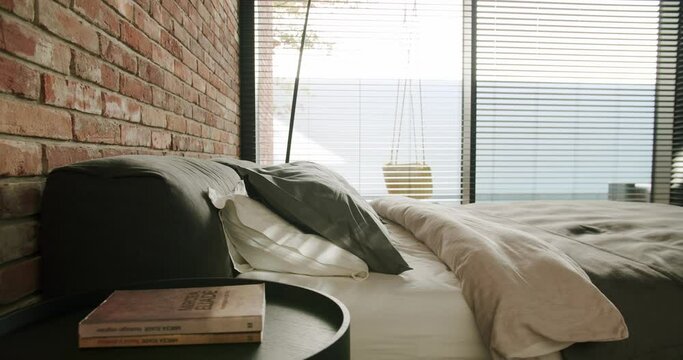 Luxury bedroom with loft interior, double bed with pillows and bedding, bedside table with books and glass wall or panoramic window with blinds. Modern modular cottage house