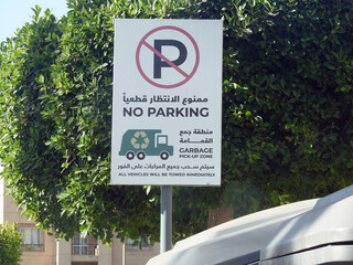 A Road sign in the street near a dumpster litter box in Arabic and English, Translation of the...