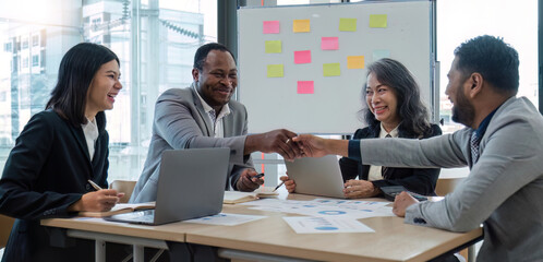 African professional businessman and Indian male successful businessman in formal suit sitting shaking hands when business agreement deal done while female colleagues clapping hands celebrating.