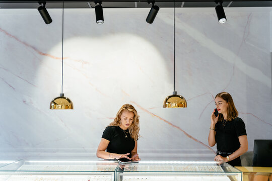 Elegant lady female ntrepreneur discussing jewelry order on smart phone at workplace her assistant working with tablet looking at the leftovers, helps to place an order.