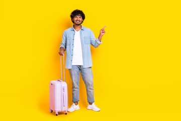 Full size photo of good mood man dressed denim shirt jeans stand with suitcase indicating empty space isolated on yellow color background