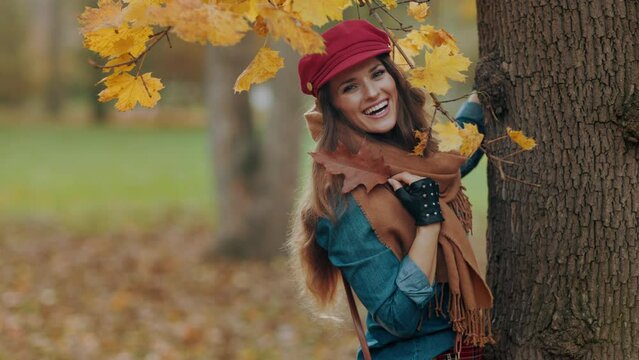 Hello autumn. happy stylish 40 years old woman in jeans shirt and red hat with autumn leaf and scarf near tree in the city park.