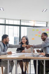 African professional businessman and Indian male successful businessman in formal suit sitting shaking hands when business agreement deal done while female colleagues clapping hands celebrating.