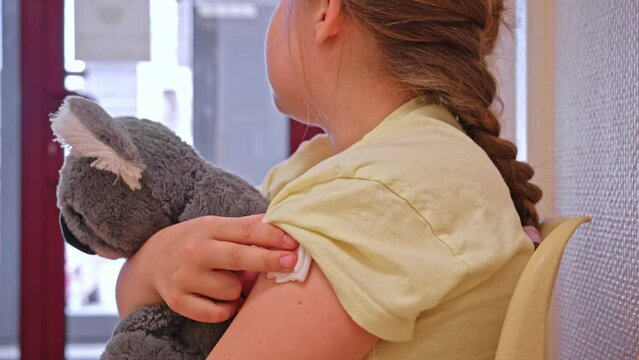 Caucasian Girl Kid Hugging Teddy Bear Sitting in Clinic Waiting Room After Vaccine Shot Pressing Gauze Pad Against Needle Injection to Stop Bleeding 
