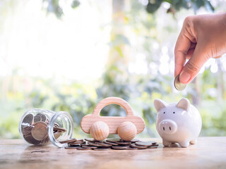 Close up of hand putting coins into piggy bank and wooden toy car on stack of coins. Transport and...